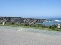 an empty road next to a cliff on a sunny day with ocean behind it and rocks, cliffs, bushes, grass