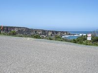 an empty road next to a cliff on a sunny day with ocean behind it and rocks, cliffs, bushes, grass