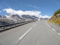 a long country road surrounded by snow capped mountains and rock sides with guard railing on both sides