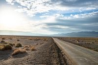 a country road in the middle of a barren field near mountains and a cloudy sky