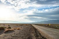 a country road in the middle of a barren field near mountains and a cloudy sky