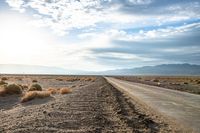 a country road in the middle of a barren field near mountains and a cloudy sky