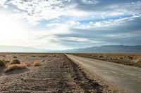 a country road in the middle of a barren field near mountains and a cloudy sky