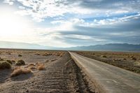 a country road in the middle of a barren field near mountains and a cloudy sky