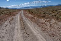 a dirt road winding through an empty field and mountain range in the distance to the right