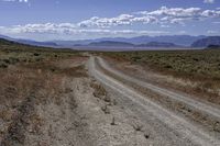 a dirt road leading to the mountains on a sunny day with white clouds and some blue sky