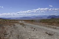 a dirt road leading to the mountains on a sunny day with white clouds and some blue sky