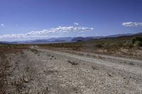 a dirt road leading to the mountains on a sunny day with white clouds and some blue sky