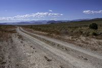 a dirt road leading to the mountains on a sunny day with white clouds and some blue sky