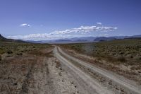 a dirt road leading to the mountains on a sunny day with white clouds and some blue sky