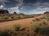 a dirt road through a barren area with tall red rocks in the distance and greenery around it