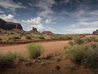 a dirt road through a barren area with tall red rocks in the distance and greenery around it