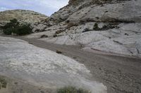 a dirt road runs in between large rocks, near trees and shrubs at the bottom of a rocky hill