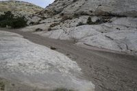 a dirt road runs in between large rocks, near trees and shrubs at the bottom of a rocky hill