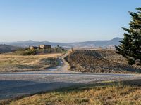 a dirt road and a tree on the edge of a mountain side with hills in the background