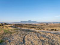 dirt road winding through an empty field towards a hill top and distant landscape with mountains in background