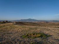 dirt road winding through an empty field towards a hill top and distant landscape with mountains in background