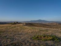 dirt road winding through an empty field towards a hill top and distant landscape with mountains in background