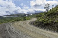 a car driving down a dirt road next to green grass and hills with a forest