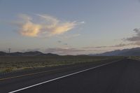 a paved desert road with a single car driving by on it in the evening with some mountains on either side