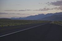 a paved desert road with a single car driving by on it in the evening with some mountains on either side