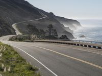 an empty highway with yellow stripes passes the ocean along a coastal trail by a steep hillside