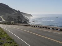 an empty highway with yellow stripes passes the ocean along a coastal trail by a steep hillside