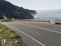 an empty highway with yellow stripes passes the ocean along a coastal trail by a steep hillside