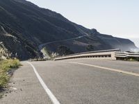 an empty highway with yellow stripes passes the ocean along a coastal trail by a steep hillside