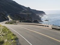 an empty highway with yellow stripes passes the ocean along a coastal trail by a steep hillside