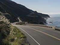 an empty highway with yellow stripes passes the ocean along a coastal trail by a steep hillside