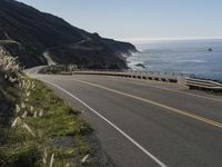 an empty highway with yellow stripes passes the ocean along a coastal trail by a steep hillside