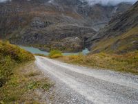 gravel road passing through a grassy valley below a mountain range with a lake, clouds, and clouds hovering behind