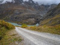 gravel road passing through a grassy valley below a mountain range with a lake, clouds, and clouds hovering behind