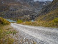 gravel road passing through a grassy valley below a mountain range with a lake, clouds, and clouds hovering behind