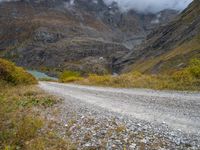 gravel road passing through a grassy valley below a mountain range with a lake, clouds, and clouds hovering behind