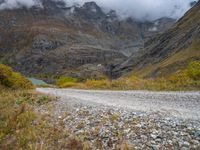 gravel road passing through a grassy valley below a mountain range with a lake, clouds, and clouds hovering behind