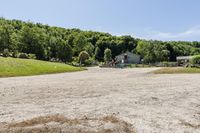 a gravel road passing through a lush green field and wooded area near a white house