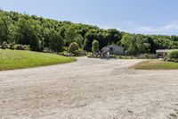 a gravel road passing through a lush green field and wooded area near a white house