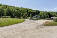 a gravel road passing through a lush green field and wooded area near a white house