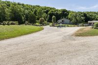 a gravel road passing through a lush green field and wooded area near a white house