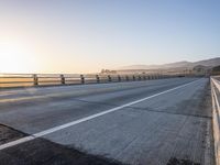 a highway leading into the distance with the sun setting in the background above it and a fence separating the roadway