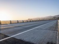 a highway leading into the distance with the sun setting in the background above it and a fence separating the roadway