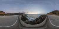 a fish - eye lens view of a scenic highway at sunrise with waves crashing under a bridge