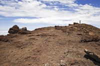 a very tall stone hill with a flag on top of it with clouds above the ground and blue skies overhead