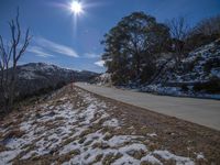 a dirt road runs along the edge of a snowy field to a rock wall and some trees