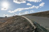 a dirt hill with an empty road and some building under the clouds and a bright sun