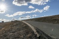 a dirt hill with an empty road and some building under the clouds and a bright sun