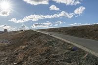 a dirt hill with an empty road and some building under the clouds and a bright sun