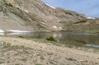 mountains around a lake are covered with snow and rocks and grass around it with small trees and shrubs in the foreground